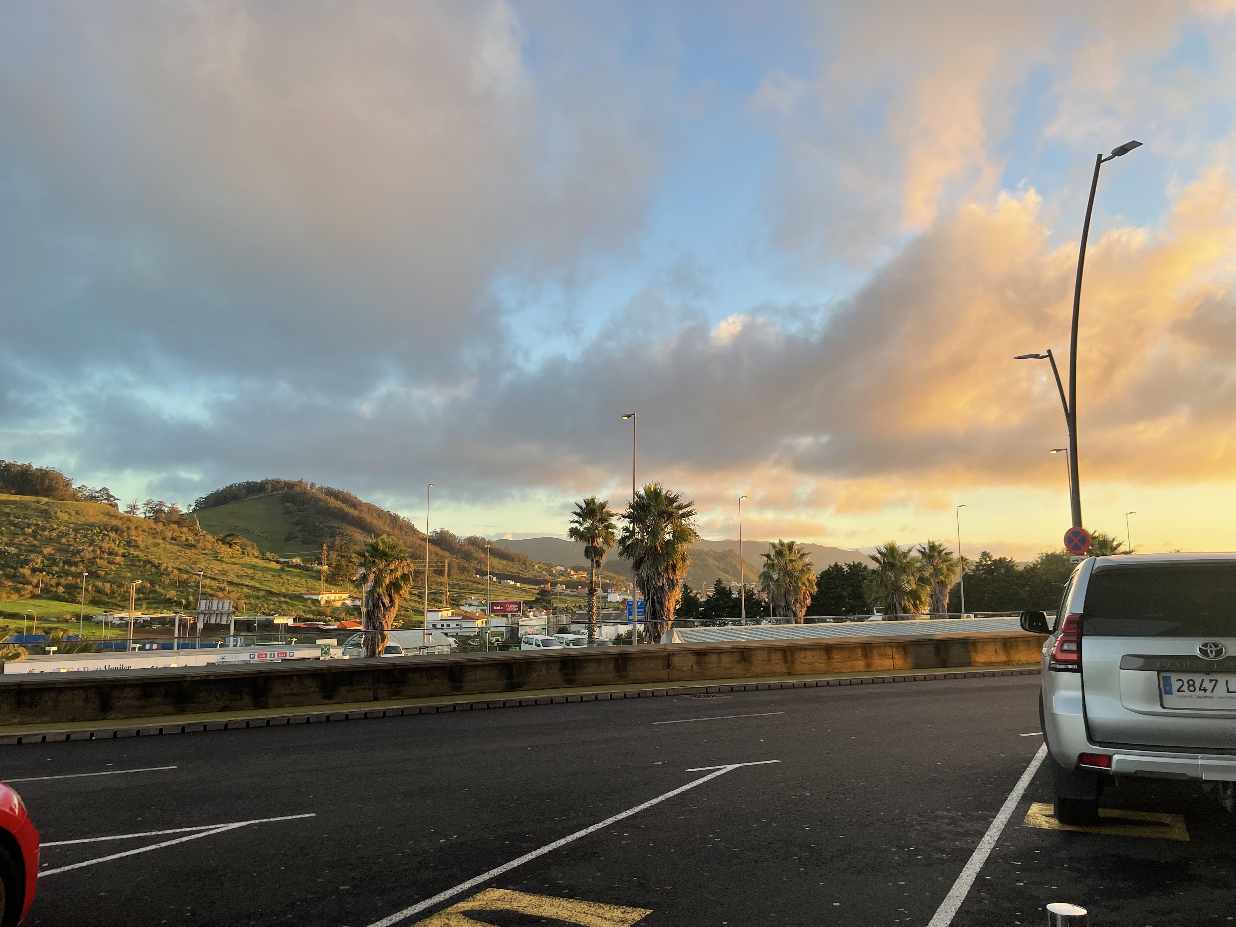 Golden hour over a parking lot with palm trees, green hills, and dramatic clouds in the Canary Islands